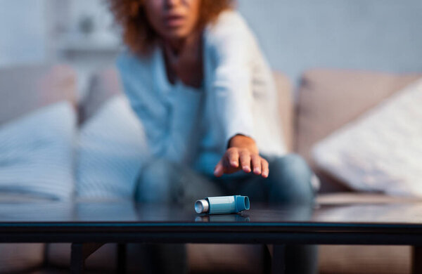 Young woman suffering asthma attack reaching inhaler, sitting on sofa at home