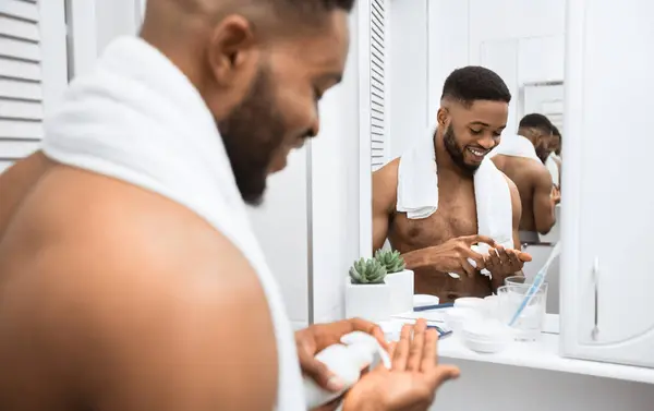 stock image Smiling afro man with naked torso squeezing cream on his hand, reflecting in mirror at bathroom. Mans skin care concept