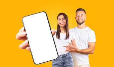 Genç bir çift boş ekranlı bir akıllı telefonu tutuyor. Adam ve kadın gülümsüyor ve kameraya bakıyor. Her ikisi de gündelik kıyafetler giyiyorlar.