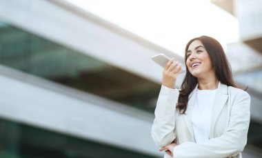 Uzun siyah saçlı genç bir kadın şehir ortamında dışarıda duruyor ve bir ses asistanı kullanırken ağzına bir akıllı telefon dayıyor.