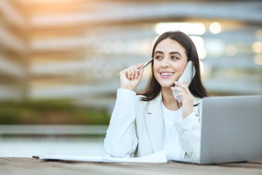 Beyaz ceketli gülümseyen genç bir iş kadını ofis binasının önünde oturuyor. Dizüstü bilgisayarı üzerinde çalışıyor ve cep telefonuyla konuşuyor. Onun önünde tahta bir masanın üzerinde kağıtlar var..