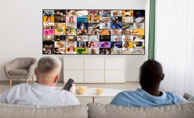 Oturma odasındaki bir kanepede oturan iki kişi büyük bir televizyon ekranını seyrediyor ve daha küçük ekranlar gösteriyor. Televizyonda gösterilen içeriğe dalmış gibi görünüyorlar.
