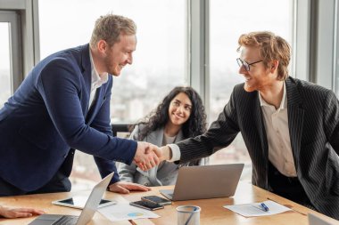 Takım elbiseli iki işadamı ofis ortamında el sıkışıyor. İkisi de gülümsüyor ve mutlu görünüyorlar. Arka planda oturan bir kadın dizüstü bilgisayarla çalışıyor..