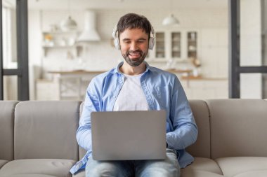 Modern bir oturma odasındaki açık gri koltukta oturan bir adam dizüstü bilgisayarı üzerinde çalışırken gülümsüyor. Kulaklık ve açık mavi düğmeli gömlek giyiyor..