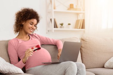 Afrikalı hamile anne, evde dizüstü bilgisayar ve kredi kartı kullanarak bebeği için kıyafet sipariş ediyor.