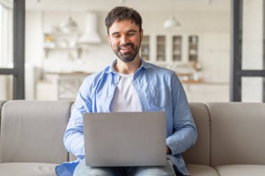 Modern bir oturma odasında açık gri bir koltukta oturan bir adam dizüstü bilgisayarıyla çalışıyor. Gülümsüyor ve işini seviyor gibi görünüyor..