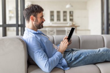 Bir adam, modern bir evde rahat bir kanepede tablet kullanarak dinleniyor. O, bacaklarını açmış ve bir yastığa yaslanmış olarak oturuyor. İçerikte kaydırıyor gibi görünüyor.