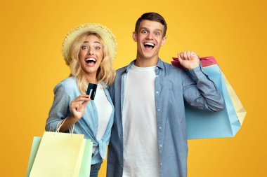 Alışveriş düşkünleri çifti. Heyecanlı genç adam ve kız ellerinde alışveriş torbaları ve sarı stüdyo arka planında kredi kartı, kopyalama alanı