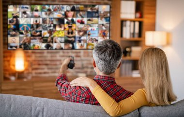 Bir çift oturma odasındaki kanepede dinleniyor. Adamın elinde uzaktan kumanda var ve daha küçük görüntüler gösteren büyük bir TV ekranına bakıyor..