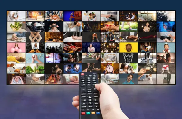 stock image A hand is holding a black remote control in front of a television screen that is displaying a variety of channels. The screen is filled with a grid of small video squares