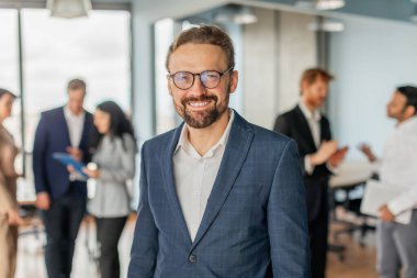 Mavi takım elbiseli ve gözlüklü bir işadamı modern ofis ortamında dururken gülümsüyor. Diğer ofis çalışanları arka planda duruyor ve konuşmalarla meşguller..