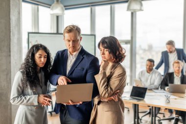 Modern bir ofiste dizüstü bilgisayara odaklanmış bir grup profesyonel iş adamı duruyor. Mavi takım elbiseli bir adam ekranda iki kadınla bilgi paylaşıyor..