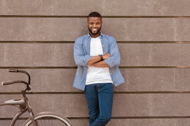 Gri şehir duvarına yaslanan, bisikletinin yanında duran ve kameraya bakan mutlu siyah adam, boşluğu kopyala.