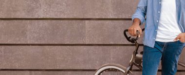 Motosikletinin yanında duran, gidonu tutan, gri şehir duvarının üzerinde poz veren tanınmayan Afrikalı adamın yakın çekimi.