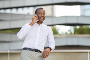 Beyaz gömlek giyen siyah bir adam balkonda güvenle duruyor ve neşeli bir telefon görüşmesi yapıyor. Çevresindeki modern mimari onun rahat tavırlarını tamamlıyor.