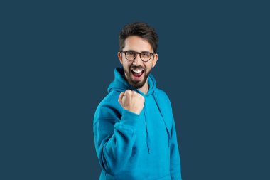 A young man wearing a bright blue hoodie expresses excitement with a joyful fist pump. His cheerful expression suggests he has just achieved something significant, and the solid background clipart