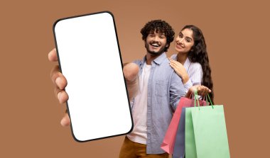 Alışveriş torbaları ve boş ekranlı akıllı telefon tutan genç bir Hintli çift. Adam telefonu kameraya doğru uzatıyor ve kadın da arkasında duruyor.