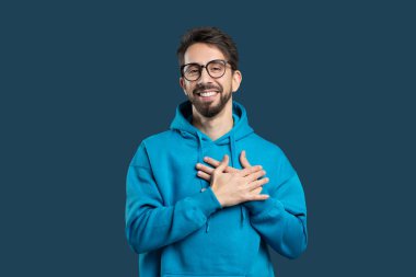 Neşeli genç bir adam mavi arka planlı, parlak mavi kapüşonlu bir stüdyoda duruyor. Ellerini kalbinin üzerine koyar. Minnettarlık ve samimiyet duygusu iletir.