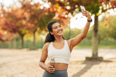 Genç bir kadın, canlı sonbahar renklerini gösteren ağaçlarla çevrili bir parkta duruyor. Akıllı telefonuyla neşeli bir selfie çekerken bir elinde su şişesi tutuyor..