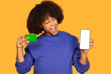 Neşeli genç bir adam bir elinde cep telefonu, diğer elinde ödeme kartıyla heyecanlı pozlar veriyor. Parlak sarı arka plan canlı atmosferi güçlendirir.