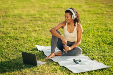 Çimenli bir parkta yoga minderinde, kulaklık takmış ve dizüstü bilgisayarla etkileşim halinde oturan bir kadın. Bir su şişesi tutuyor ve sanal egzersiz sınıfına katılırken gülümsüyor.