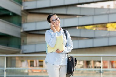 Resmi giyinmiş Asyalı kadın dışarıda duruyor, telefonda konuşurken gülümsüyor. Bir elinde evrakları tutarken diğer kolu da yanında gevşiyor, boşluğu kopyala.