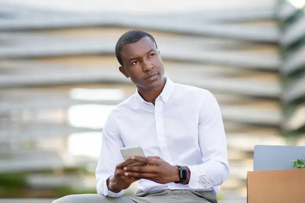 stock image A young black man, dressed in a smart white shirt, is sitting comfortably in a contemporary urban plaza. He is focused on his smartphone, presumably communicating or accessing information