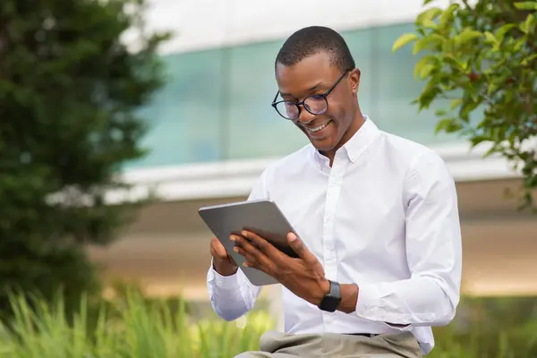 stock image African American young man wearing glasses sits comfortably in a lush outdoor setting, focused on a tablet device. He appears to be enjoying the moment, surrounded by vibrant greenery