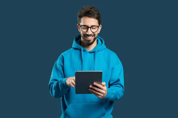 stock image A young man with glasses and a cheerful smile is actively using a tablet device in an indoor environment, dressed in a comfortable blue hoodie. His focused expression reflects engagement