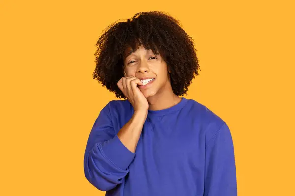 stock image A young man with curly hair is nervously biting his nails while wearing a blue shirt. The bright yellow background enhances the emotional expression, capturing a moment of anxiety or uncertainty