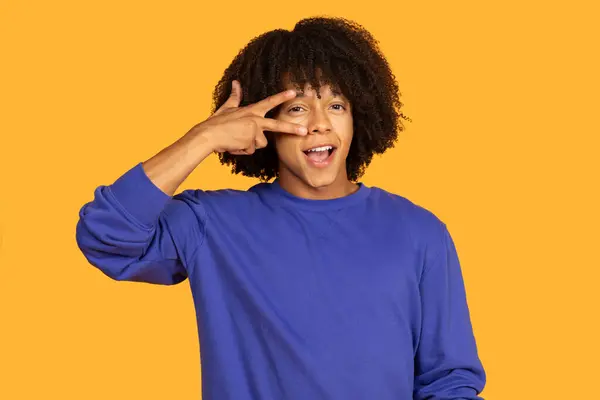 stock image A cheerful young man smiles while playfully posing with a peace sign. Their curly hair and vibrant blue attire contrast beautifully with the bright yellow backdrop, creating a lively atmosphere.