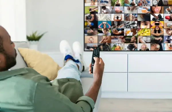 stock image Black man is relaxing on a couch in his living room, watching television with a remote control in his hand. He is wearing casual clothing and has his feet up on the couch.