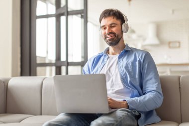 Modern bir oturma odasında beyaz bir koltukta oturan bir adam, kulaklık takıyor ve dizüstü bilgisayar kullanırken gülümsüyor. Çevrimiçi zaman geçirmekten hoşlanıyor gibi görünüyor, muhtemelen bir film izliyor, bir proje üzerinde çalışıyor..