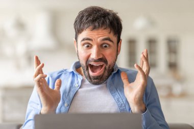 Şaşırmış bir ifadeye sahip bir adam dizüstü bilgisayarın önünde oturuyor, elleri heyecanla ekrana bakarken.