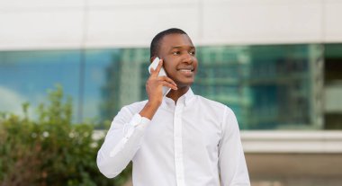 Beyaz gömlek giyen neşeli bir siyah adam dışarıda duruyor, hoş bir ifadeyle telefonuyla konuşuyor. Arka planda modern mimari ve yeşillik yer alıyor.