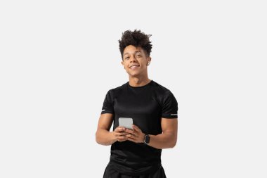 Genç bir adam, elinde akıllı telefonu tutarken sırıtarak güvenle ayakta duruyor. O, siyah bir atletik gömlek ve şort giydi. Parlak iç mekan onun neşeli tavrını geliştirir..