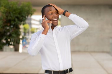 Genç siyahi bir adam modern bir binanın yanında duruyor, telefonuyla konuşuyor ve neşe ve heyecan ifade ediyor..