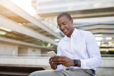 Genç siyah bir adam rahat bir şekilde oturur, akıllı telefonuna odaklanır ve modern bir kentsel bölgenin canlı atmosferinin tadını çıkarır. Sıcak güneş ışığı neşeli çevreyi güçlendirir..