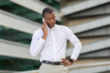 Genç bir siyah adam akıllı telefonuyla konuşurken modern bir şehir ortamında düşünceli bir şekilde duruyor. Gıcır gıcır beyaz bir gömlek ve bej pantolon giyiyor..