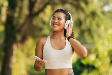 Kıvırcık saçlı bir kadın büyük kulaklık takarken neşeyle gülümsüyor. Bir elinde akıllı bir telefon tutuyor ve yemyeşil bir parkta çalışmaktan zevk alıyor gibi görünüyor.