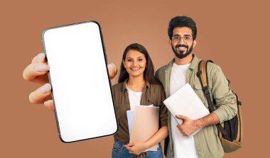 İki gülümseyen Hintli öğrenci kahverengi bir arka planın önünde duruyor. Kitapları ve boş ekranlı bir akıllı telefonu tutuyorlar.