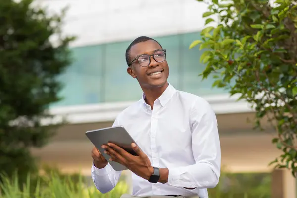 stock image A young black man dressed in a white shirt stands outdoors, happily engaging with a tablet. He appears to be in a lush urban environment, surrounded by greenery and modern architecture.