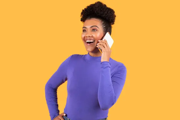 stock image A cheerful young woman engages in a lively conversation on her smartphone, smiling widely. She stands with one hand on her hip, exuding confidence and happiness against a vibrant yellow backdrop