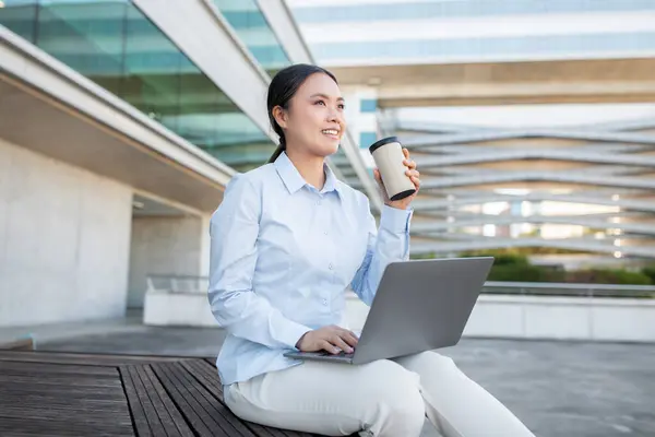 stock image A young Asian professional sits outside a contemporary office building, typing on a laptop. She holds a cup of coffee, appearing focused and relaxed in her workspace, copy space