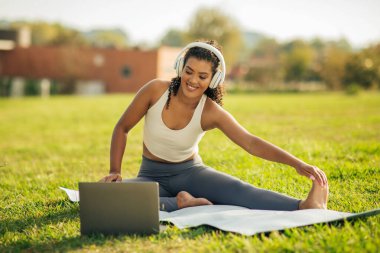 Genç bir kadın çimenli bir tarlada yoga yaparak güneşli bir günün tadını çıkarıyor. Bacağını esnetirken kulaklık takıyor. Huzurlu bir ortamda zihni ve vücuduyla bağlantı kuruyor..