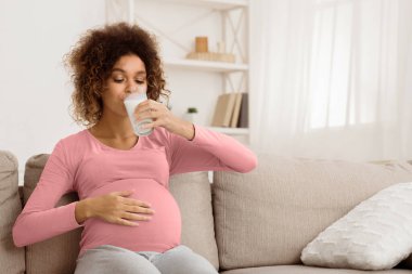 Hamilelik için faydalı bir diyet. Afro hamile kadın süt içiyor, kanepede dinleniyor, boş yer var.