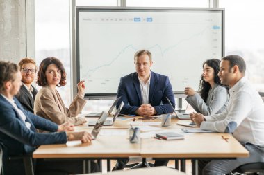 Bir grup iş adamı modern bir ofiste masanın etrafında toplanarak büyük ekranda gösterilen bir büyüme tablosunu tartışıyor. Takım hareketli bir sohbete daldı.