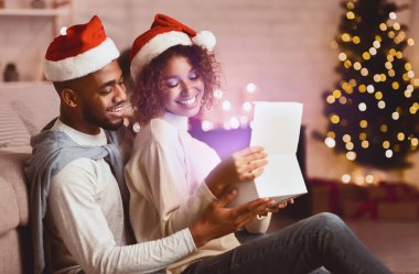 Mutlu afro çift Noel hediyesi kutusunu oturma odasında, Xmas ağacının önünde açıyor.
