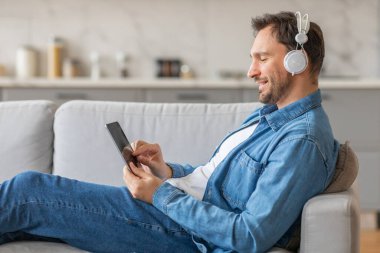 Bir adam açık gri bir koltukta rahatlıyor, kot pantolon ve mavi kot gömlek giyiyor, siyah bir tablete bakıyor. O, beyaz kulaklık takarken müzik dinlerken gülümsüyor..