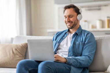 Oturma odasındaki beyaz koltukta oturan bir adam, kulaklık takıyor ve dizüstü bilgisayarla çalışıyor. Gülümsüyor, işinden zevk aldığını ya da müzik dinlediğini gösteriyor..